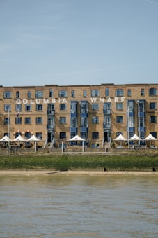 A large brick building with multiple windows and blue accents stands behind a waterfront area. The structure has the text 'COLUMBIA WHARF' prominently displayed on its facade. In front of the building, there is a row of white triangular tents and a grassy area leading down to the water.