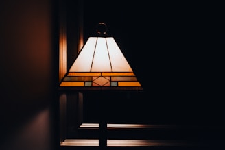 A stained glass lampshade emits a warm glow against a dark background. The design includes geometric patterns with shades of amber, yellow, and green, creating an artistic and vintage aesthetic.