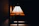 A stained glass lampshade emits a warm glow against a dark background. The design includes geometric patterns with shades of amber, yellow, and green, creating an artistic and vintage aesthetic.