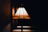 A stained glass lampshade emits a warm glow against a dark background. The design includes geometric patterns with shades of amber, yellow, and green, creating an artistic and vintage aesthetic.