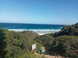 A scenic view of a beach with vibrant blue ocean waves meeting the sandy shore. Lush green foliage surrounds a pathway leading to the beach, with a recycling bin and a wooden sign visible near the entrance.