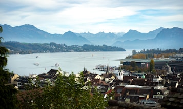 The image features a picturesque view of a lakeside town with a large body of water surrounded by mountains in the background. Several boats are visible on the lake, and the shoreline is dotted with buildings and trees, showcasing a mix of urban and natural scenery. The sky is cloudy with patches of blue, contributing to a serene and tranquil atmosphere.
