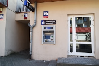 An outdoor ATM is mounted on the wall of a building. The machine is labeled with a sign that reads 'bankomat' offering 24-hour service. Nearby are a drainpipe and a doorway with a glass door that leads inside the building. The building has a beige facade and there are signs with the numbers 14a and 14b displayed.