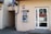 An outdoor ATM is mounted on the wall of a building. The machine is labeled with a sign that reads 'bankomat' offering 24-hour service. Nearby are a drainpipe and a doorway with a glass door that leads inside the building. The building has a beige facade and there are signs with the numbers 14a and 14b displayed.