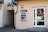 An outdoor ATM is mounted on the wall of a building. The machine is labeled with a sign that reads 'bankomat' offering 24-hour service. Nearby are a drainpipe and a doorway with a glass door that leads inside the building. The building has a beige facade and there are signs with the numbers 14a and 14b displayed.