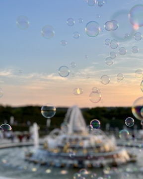 Soap bubbles float in the air against a soft sunset sky, with a fountain blurred in the background. The bubbles catch the light, displaying a spectrum of colors.