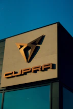 A building facade prominently displays the Cupra logo in metallic tones against a clear blue sky. The sleek, angular design of the logo is positioned above the brand name, also in metallic letters.