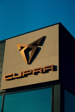A building facade prominently displays the Cupra logo in metallic tones against a clear blue sky. The sleek, angular design of the logo is positioned above the brand name, also in metallic letters.