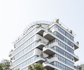 A modern, multi-story building with rounded edges and extensive glass windows. The structure features several terraces with plants, giving it a green, eco-friendly appearance. It is designed with clean, smooth lines and a contemporary architectural style.