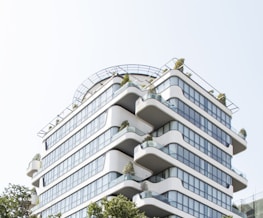 A modern, multi-story building with rounded edges and extensive glass windows. The structure features several terraces with plants, giving it a green, eco-friendly appearance. It is designed with clean, smooth lines and a contemporary architectural style.