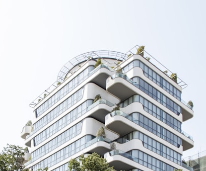 A modern, multi-story building with rounded edges and extensive glass windows. The structure features several terraces with plants, giving it a green, eco-friendly appearance. It is designed with clean, smooth lines and a contemporary architectural style.