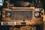 Front view of a rugged Land Rover Defender covered in dirt and mud, featuring a spare tire on the hood. The British flag decal is present on the right, and the vehicle is illuminated by warm, natural sunlight, enhancing its earthy tones.