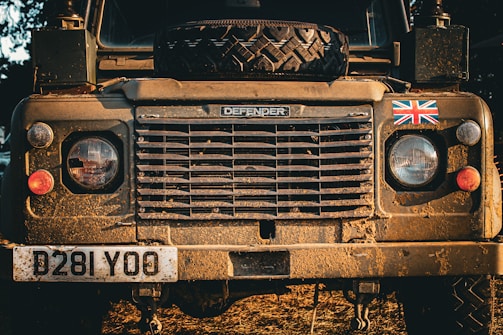 Front view of a rugged Land Rover Defender covered in dirt and mud, featuring a spare tire on the hood. The British flag decal is present on the right, and the vehicle is illuminated by warm, natural sunlight, enhancing its earthy tones.