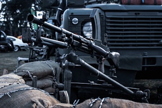 A mounted machine gun is positioned on a military vehicle. The setup includes sandbags and barbed wire in the foreground. The vehicle has a rugged design, typical of military transport, with details like a round headlamp and a grill.