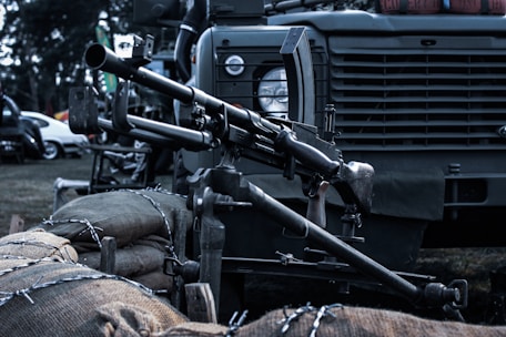 A mounted machine gun is positioned on a military vehicle. The setup includes sandbags and barbed wire in the foreground. The vehicle has a rugged design, typical of military transport, with details like a round headlamp and a grill.