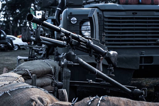 A mounted machine gun is positioned on a military vehicle. The setup includes sandbags and barbed wire in the foreground. The vehicle has a rugged design, typical of military transport, with details like a round headlamp and a grill.