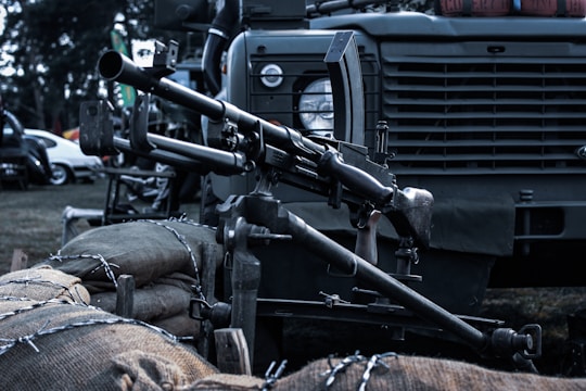 A mounted machine gun is positioned on a military vehicle. The setup includes sandbags and barbed wire in the foreground. The vehicle has a rugged design, typical of military transport, with details like a round headlamp and a grill.