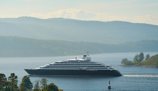 A large, luxurious yacht sails on a calm body of water surrounded by lush, green hills and forests. The backdrop includes layers of misty hills under a partly cloudy sky, creating a serene and tranquil scene.