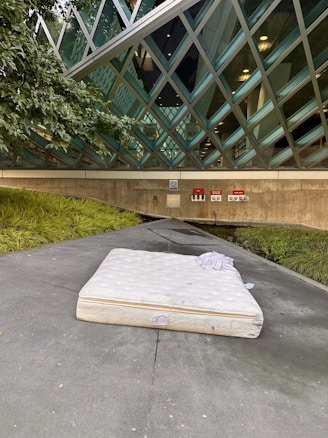 A mattress lies on a concrete path in front of a building with a modern architectural style featuring large, diamond-shaped glass panels. There are green plants on the left side, and the building's reflective glass reveals some interior lights. The ground is slightly wet, and a piece of cloth rests on top of the mattress.