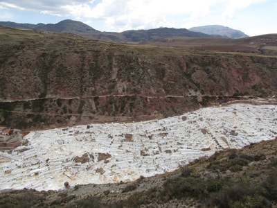 A mountainous landscape featuring a large, terraced area filled with white rectangular pools. The terraces are situated in a valley and are used for salt mining. The surrounding area consists of green and brown vegetation with distant mountains under a partly cloudy sky.