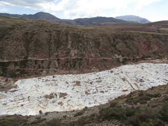 A mountainous landscape featuring a large, terraced area filled with white rectangular pools. The terraces are situated in a valley and are used for salt mining. The surrounding area consists of green and brown vegetation with distant mountains under a partly cloudy sky.