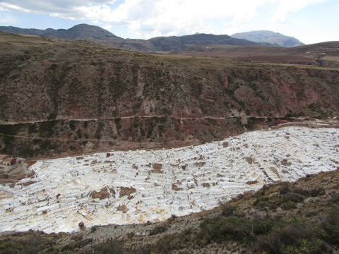 A mountainous landscape featuring a large, terraced area filled with white rectangular pools. The terraces are situated in a valley and are used for salt mining. The surrounding area consists of green and brown vegetation with distant mountains under a partly cloudy sky.