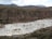 A mountainous landscape featuring a large, terraced area filled with white rectangular pools. The terraces are situated in a valley and are used for salt mining. The surrounding area consists of green and brown vegetation with distant mountains under a partly cloudy sky.