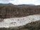 A mountainous landscape featuring a large, terraced area filled with white rectangular pools. The terraces are situated in a valley and are used for salt mining. The surrounding area consists of green and brown vegetation with distant mountains under a partly cloudy sky.