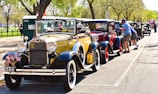A row of vintage cars is parked alongside a street, with people interacting with the vehicles. The cars are from different decades, primarily focusing on classic models like Ford Model A. Flags and decorative elements are visible on the cars, and the setting is outdoors with trees and green foliage around.