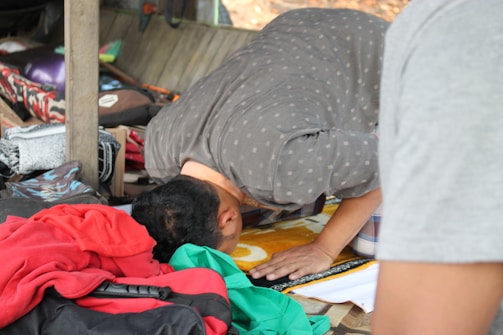 A person is seen bending forward in a position associated with prayer or meditation. They are surrounded by an assortment of colorful clothes and personal items in what appears to be a compact space. The setting suggests a sense of improvisation or temporary arrangement.