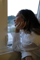 Calm woman sitting thoughtfully by a window with soft charcoal curtains, lost in reflection.