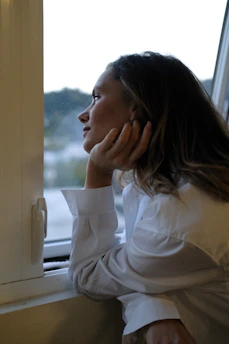 Calm woman sitting thoughtfully by a window with soft charcoal curtains, lost in reflection.
