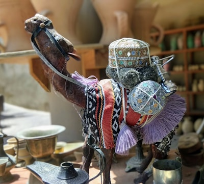 A decorative camel figurine adorned with colorful, intricate textiles and beadwork. The camel is placed among various metallic and ceramic objects on a table, with a blurred background of pottery and crafts.