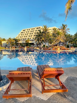 A serene resort setting with two empty lounge chairs overlooking a reflective swimming pool. The pool reflects the surrounding palm trees and a large, modern hotel building. The scene is bathed in warm sunlight, suggesting a tropical or vacation destination.