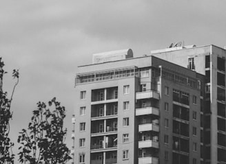 A high-rise residential building with multiple balconies and large windows. The structure appears to have a modern design, with a flat roof and some satellite dishes visible on top. The sky is overcast, and there are tree branches in the foreground.