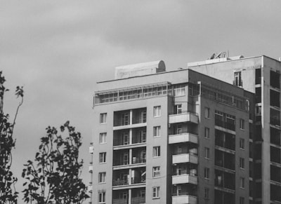 A high-rise residential building with multiple balconies and large windows. The structure appears to have a modern design, with a flat roof and some satellite dishes visible on top. The sky is overcast, and there are tree branches in the foreground.