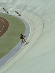 Cyclists are racing on an indoor velodrome, each rider leaning into the curved track. The scene captures the dynamic motion of the sport, with shadows cast by the riders enhancing the sense of speed.