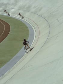 Cyclists are racing on an indoor velodrome, each rider leaning into the curved track. The scene captures the dynamic motion of the sport, with shadows cast by the riders enhancing the sense of speed.