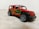 A detailed die-cast model of a red SUV with large black wheels and graphic designs on the side, including green and orange accents. The vehicle is positioned on a softly textured surface with a blurred background.