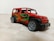 A detailed die-cast model of a red SUV with large black wheels and graphic designs on the side, including green and orange accents. The vehicle is positioned on a softly textured surface with a blurred background.