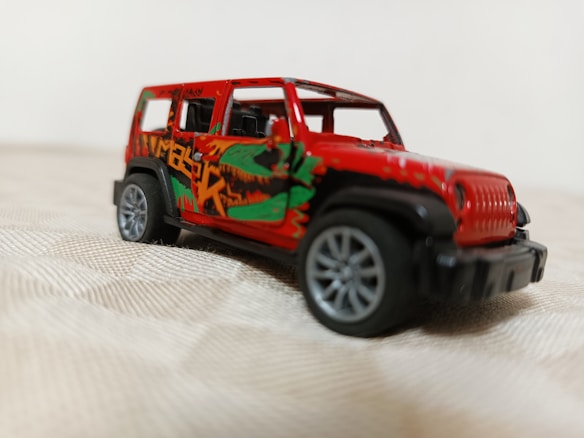 A detailed die-cast model of a red SUV with large black wheels and graphic designs on the side, including green and orange accents. The vehicle is positioned on a softly textured surface with a blurred background.