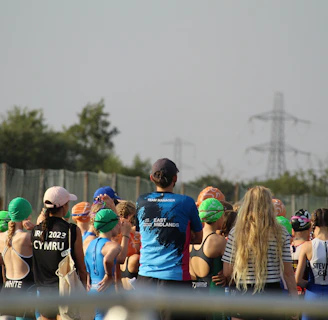A group of people, mostly young individuals, stand together in what appears to be a sporting event. They are wearing swim caps of various colors, including green, orange, and blue, and some have sports jerseys with text on the back. There are trees and electricity pylons in the background, indicating an outdoor setting.