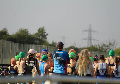 A group of people, mostly young individuals, stand together in what appears to be a sporting event. They are wearing swim caps of various colors, including green, orange, and blue, and some have sports jerseys with text on the back. There are trees and electricity pylons in the background, indicating an outdoor setting.