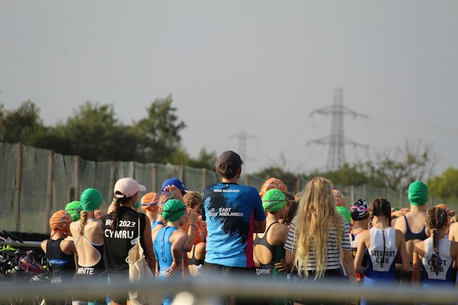 A group of people, mostly young individuals, stand together in what appears to be a sporting event. They are wearing swim caps of various colors, including green, orange, and blue, and some have sports jerseys with text on the back. There are trees and electricity pylons in the background, indicating an outdoor setting.