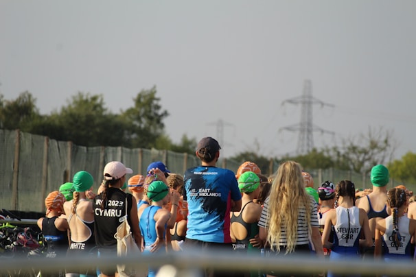 A group of people, mostly young individuals, stand together in what appears to be a sporting event. They are wearing swim caps of various colors, including green, orange, and blue, and some have sports jerseys with text on the back. There are trees and electricity pylons in the background, indicating an outdoor setting.