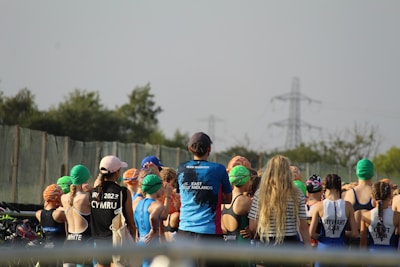 A group of people, mostly young individuals, stand together in what appears to be a sporting event. They are wearing swim caps of various colors, including green, orange, and blue, and some have sports jerseys with text on the back. There are trees and electricity pylons in the background, indicating an outdoor setting.