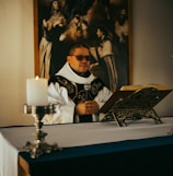 A person dressed in religious attire is standing behind an altar, holding a microphone. A lit candle is on a decorative holder at the foreground, and an open book rests on an ornate book stand. A large painting with religious imagery hangs in the background, depicting several figures in dramatic poses.