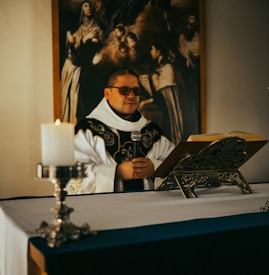 A person dressed in religious attire is standing behind an altar, holding a microphone. A lit candle is on a decorative holder at the foreground, and an open book rests on an ornate book stand. A large painting with religious imagery hangs in the background, depicting several figures in dramatic poses.