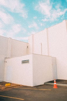 A minimalistic architectural scene featuring white building structures under a bright blue sky. The geometric shapes of the buildings create a clean and modern aesthetic. A single orange traffic cone is placed on the ground near the corner of the building, adding a pop of color.