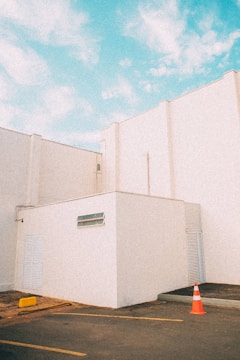 A minimalistic architectural scene featuring white building structures under a bright blue sky. The geometric shapes of the buildings create a clean and modern aesthetic. A single orange traffic cone is placed on the ground near the corner of the building, adding a pop of color.